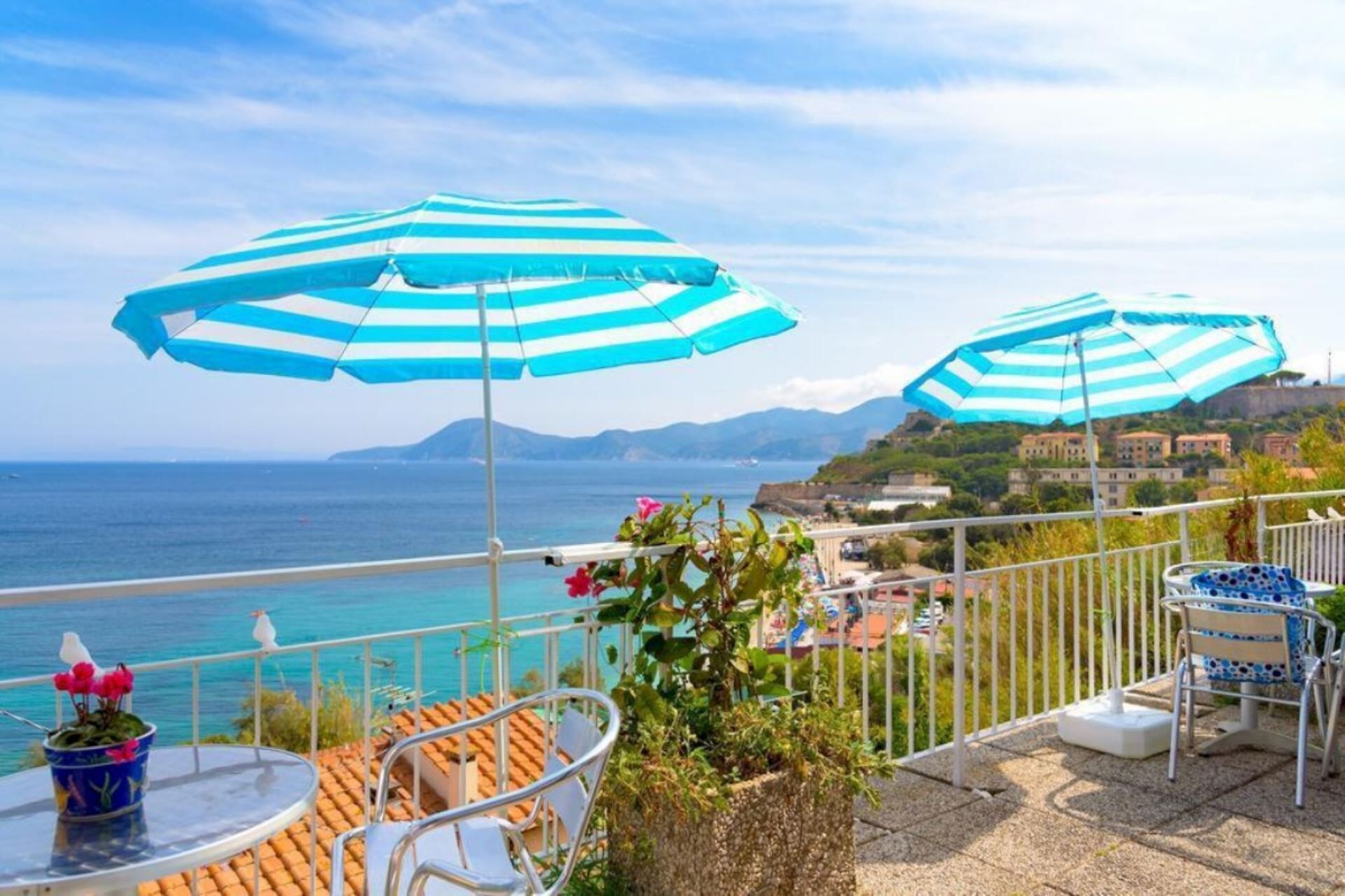 A balcony with two striped umbrellas, a potted plant, and a view of the sea and buildings.