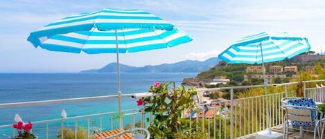 A balcony with two striped umbrellas, a potted plant, and a view of the sea and buildings.