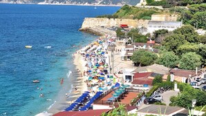 A coastal town with a beach, a fortified structure, and a mountainous backdrop.