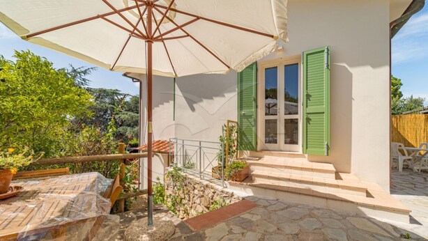 A stone patio with a table and chairs, a covered area with an umbrella, and a building with green shutters.