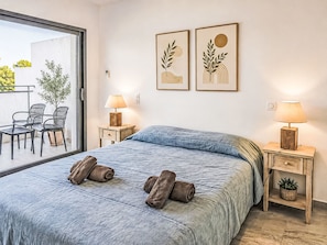 A bedroom with a bed, two bedside tables with lamps, a window with a view of outdoor furniture, and two wall-mounted artworks.