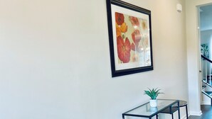 A modern dining area with a glass table and two black metal stools. A framed floral artwork hangs on the wall above the table. - Newly constructed 6 bedroom home (Frederick)