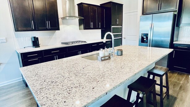 A modern kitchen with dark cabinets, a large island with stools, a stainless steel refrigerator, and a built-in oven. - Newly constructed 6 bedroom home (Frederick)