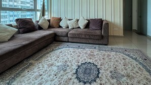 Een woonkamer met een hoekbank, een kamerbreed tapijt en uitzicht op een gebouw door het raam.