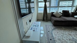 Een woonkamer met een wandgemonteerde flatscreen televisie, een bank met kussens, een kamerkleed met een patroon en een raam met gordijnen.
