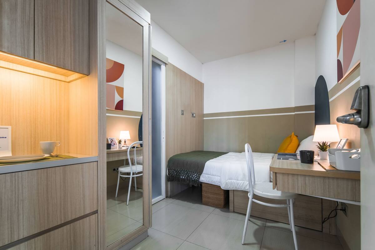 A compact hotel room with a bed, a desk, a chair, a small table, and a mirror.