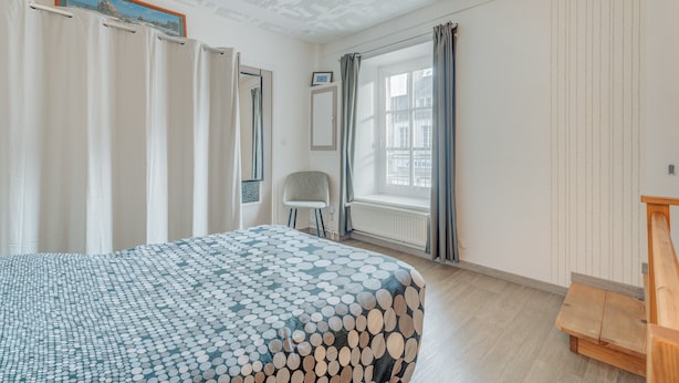 Un dormitorio con cama, una silla, un banco de madera, una ventana con cortinas, un radiador y un ventilador de techo.