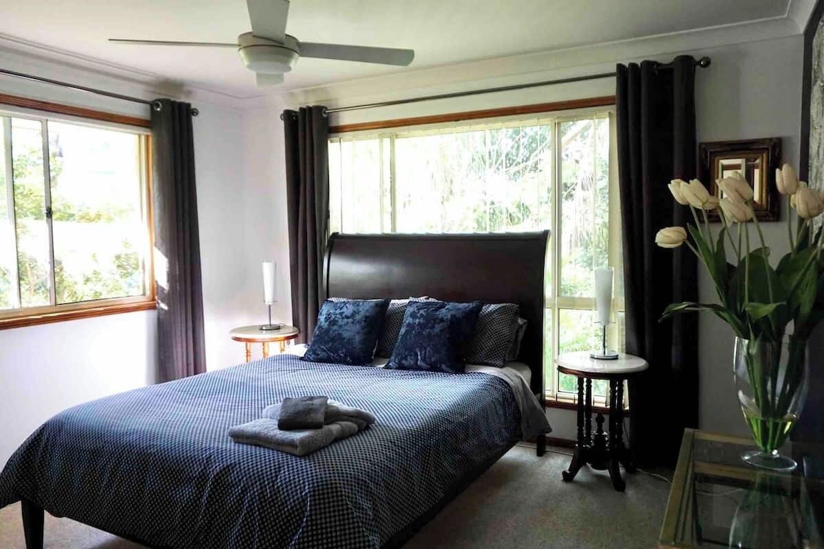 Une chambre à coucher avec un grand lit, un ventilateur de plafond, une table de chevet, un vase avec des fleurs et une fenêtre avec des rideaux.