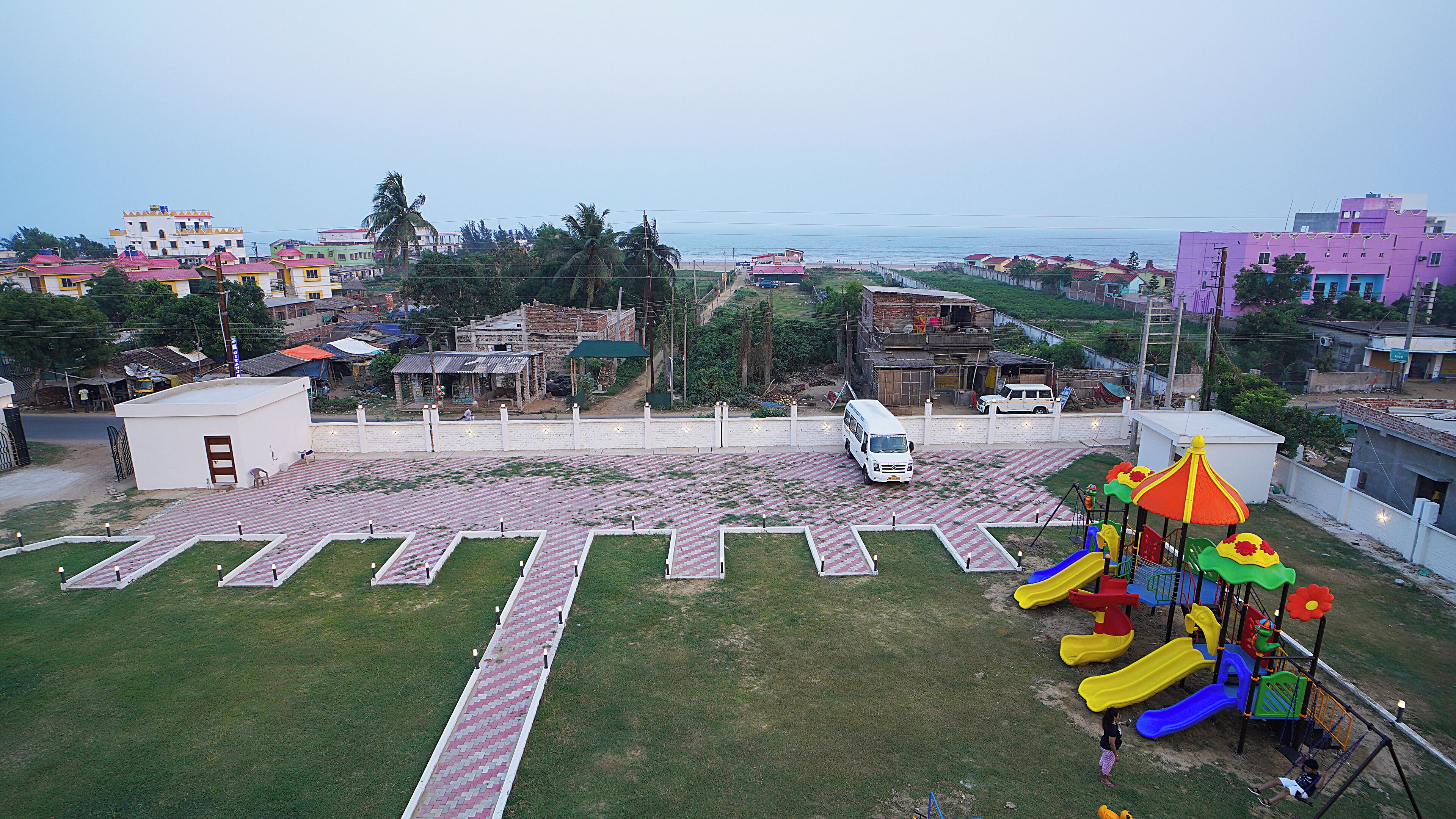 A playground with a slide and swings, a white van parked nearby, and a view of a beach and residential buildings in the background.
