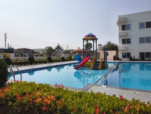 En swimmingpool med en vattenrutschkana och en lekplats för barn, omgiven av grönska och med en byggnad i bakgrunden.
