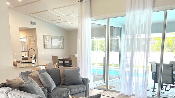 Un salon moderne avec un canapé d’angle gris, un tapis à motifs et une vue sur une piscine, visible à travers de grandes portes vitrées.