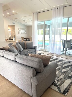 Un salon moderne avec un canapé d’angle gris, un tapis à motifs et une vue sur une piscine, visible à travers de grandes portes vitrées.