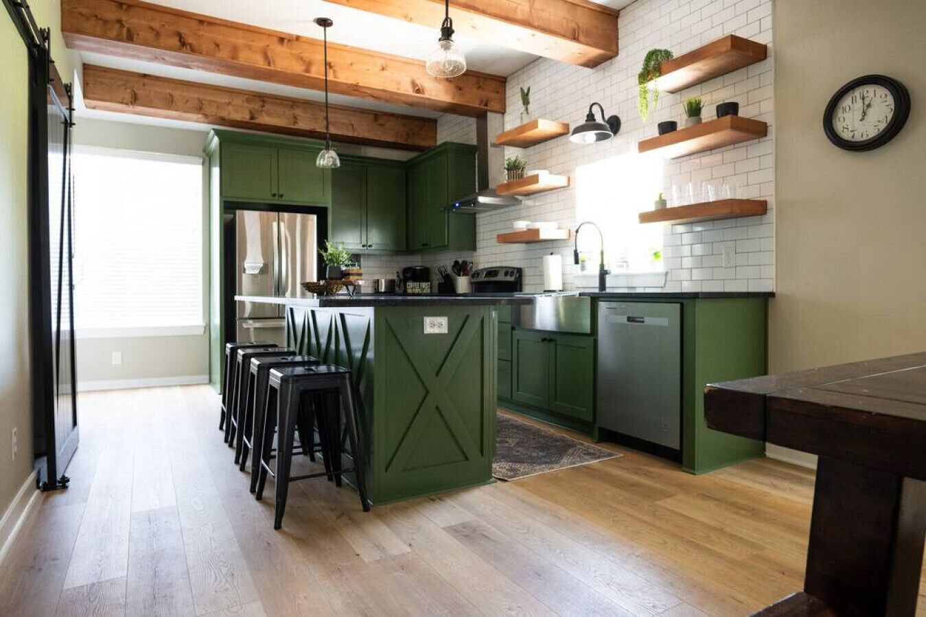 A kitchen with green cabinets, wooden beams, and a dining area with stools.