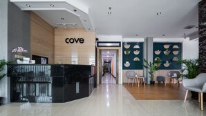 A modern hotel lobby with a reception desk, a hallway leading to rooms, and a seating area with plants and chairs. - Cove Taman (Jakarta)