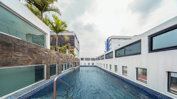 A swimming pool with a wooden deck, surrounded by modern buildings with large windows and a stone wall. - Cove Taman (Jakarta)