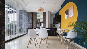 A modern dining area with white chairs and a round table, large windows, and a yellow accent wall. - Cove Taman (Jakarta)