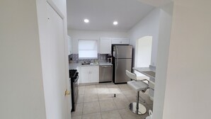 A compact kitchen with white cabinets, a stainless steel refrigerator, a sink, and a small bar stool.