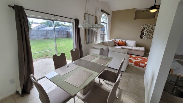 A dining area with a glass table and chairs, a sofa in the background, and a view of the backyard through large windows.