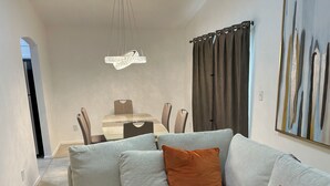 A modern living room with a grey sofa, a remote control, and a dining area with a table and chairs.