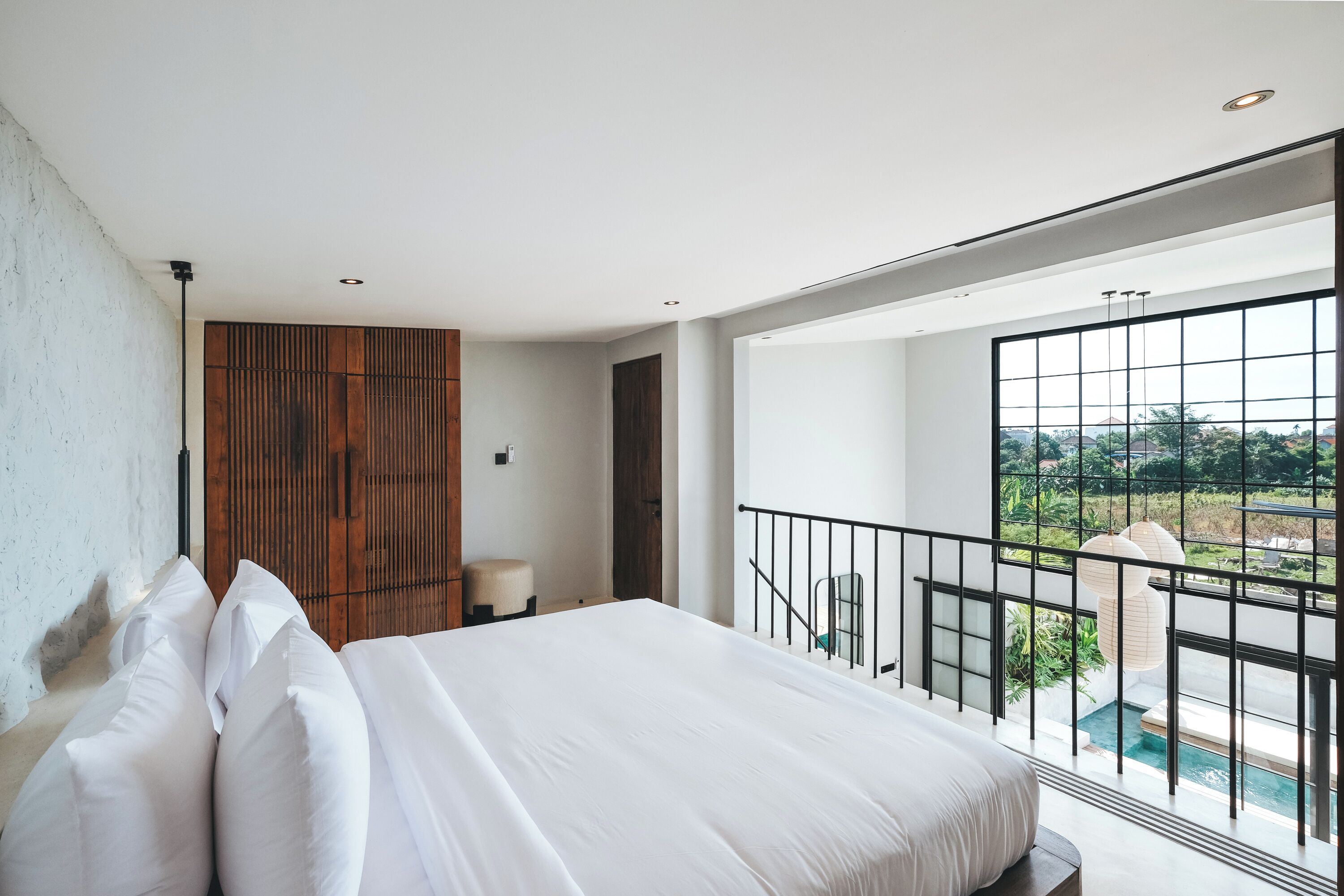 Ein modernes Schlafzimmer mit einem großen Bett, einer Wandpaneele aus Holz, einem Balkon mit Blick auf Grünflächen und einem Fenster mit Poolblick.