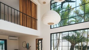 Amplia sala de estar con un sofá blanco curvo, mesas de centro de madera y un ventanal con vistas a palmeras.