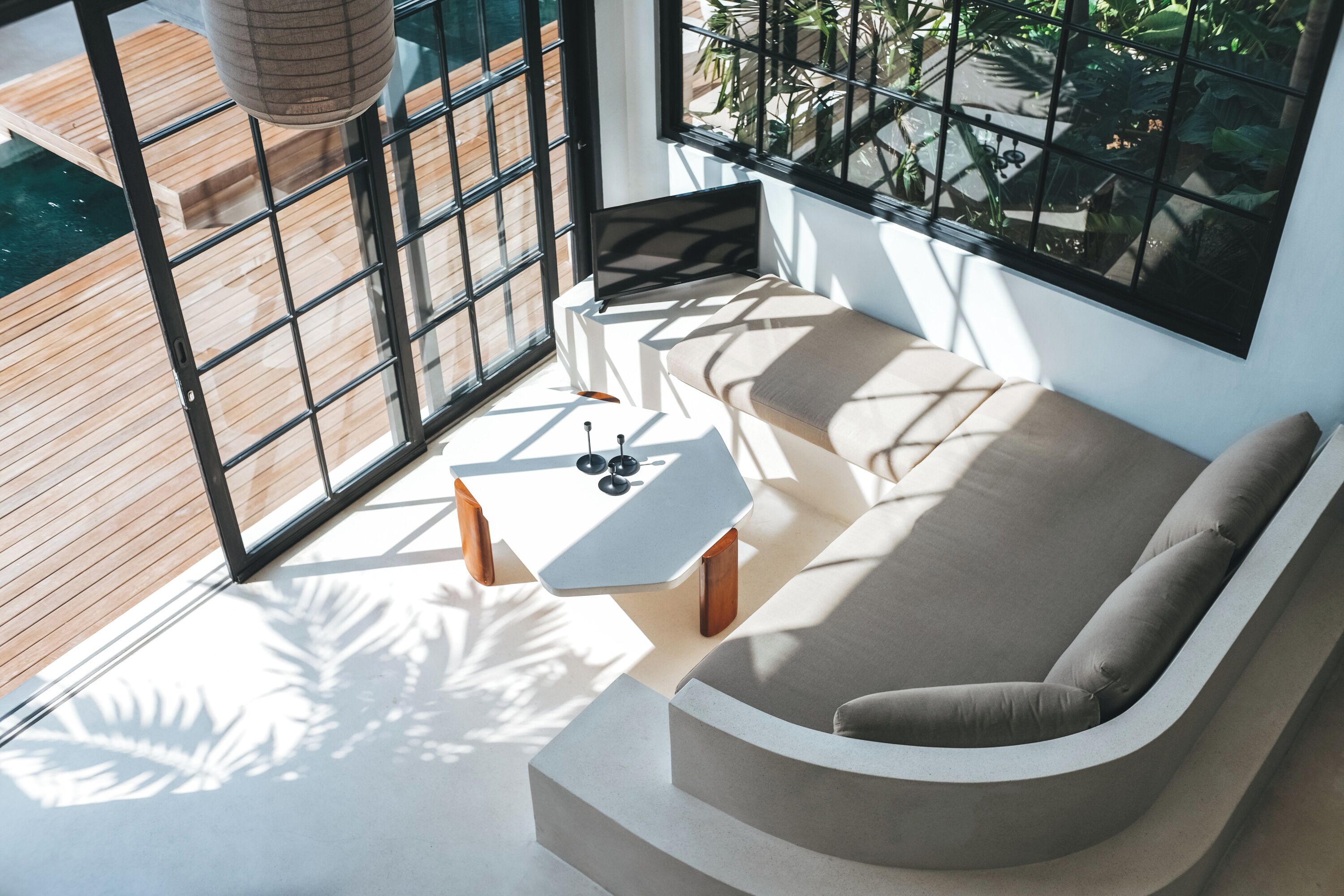 Et moderne interiør med en buet sofa, et lille hvidt bord og et stort vindue med udsigt til en træterrasse og grøn vegetation.