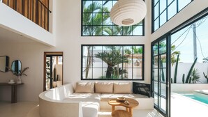 Una sala de estar moderna con un sofá curvo, una mesa de centro de madera y ventanas amplias con vistas a una piscina y palmeras.