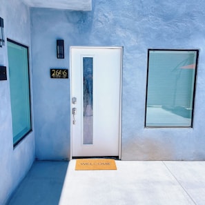 Una puerta con un letrero que dice '2606 Main Street' encima, un felpudo de bienvenida al frente y una ventana con persianas a la derecha.