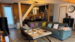 A modern living room with a grey sofa, wooden coffee table, and a kitchen area with a refrigerator and wooden cabinets.