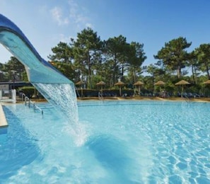 A swimming pool with a water slide, surrounded by trees and umbrellas.