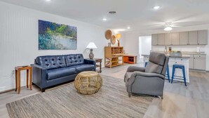 A modern living room with a blue sofa, a grey armchair, and a round wicker coffee table. A painting of a landscape is on the wall, and there is a kitchen area in the background. - Eagle's Nest - Upscale Loft,Downtown,Close to GSU! (Statesboro)