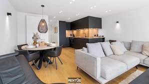 Ein modernes Wohnzimmer mit grauem Sofa, einem Esstisch mit Stühlen und einer Küchenzeile mit dunklen Schränken und einer Holz Arbeitsplatte.