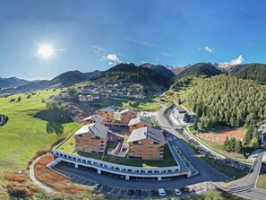 A modern building with a central courtyard, surrounded by parking and roads, set in a mountainous area with greenery and distant hills.