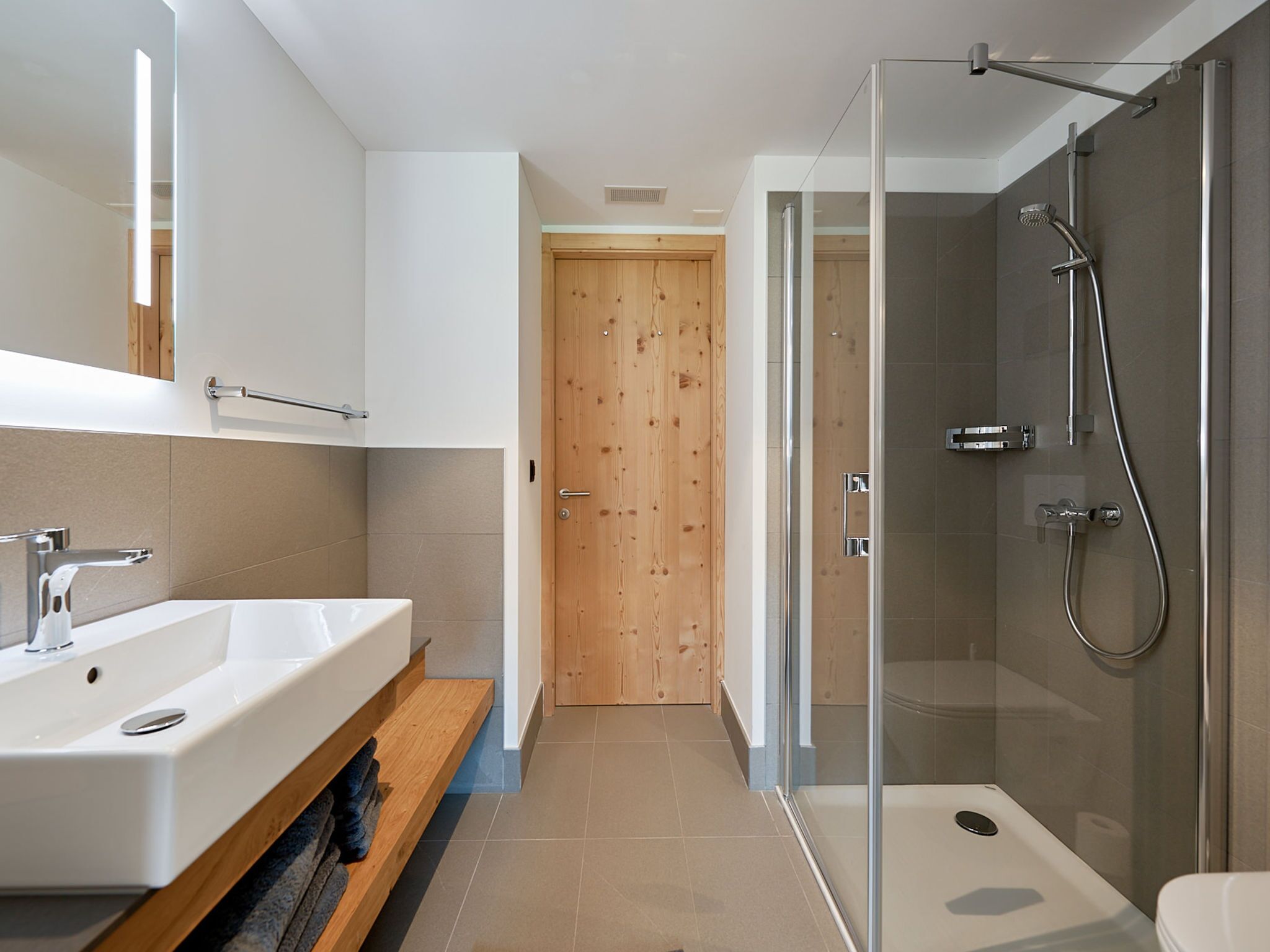 A modern bathroom with a glass shower enclosure, a wooden door, and a vanity with a sink and towel rack.