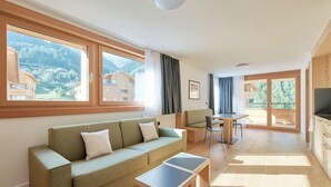 A modern living room with a sofa, a coffee table, and a dining area with a table and chairs. Large windows offer a view of the surrounding buildings and landscape.
