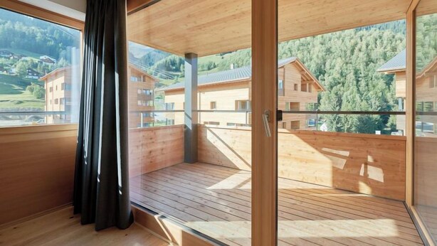 A spacious balcony with wooden flooring and large windows offering a view of a mountainous landscape.