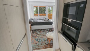 Een compacte keuken met een inbouwoven en magnetron, een klein krukje en een raam met uitzicht op een balkon.