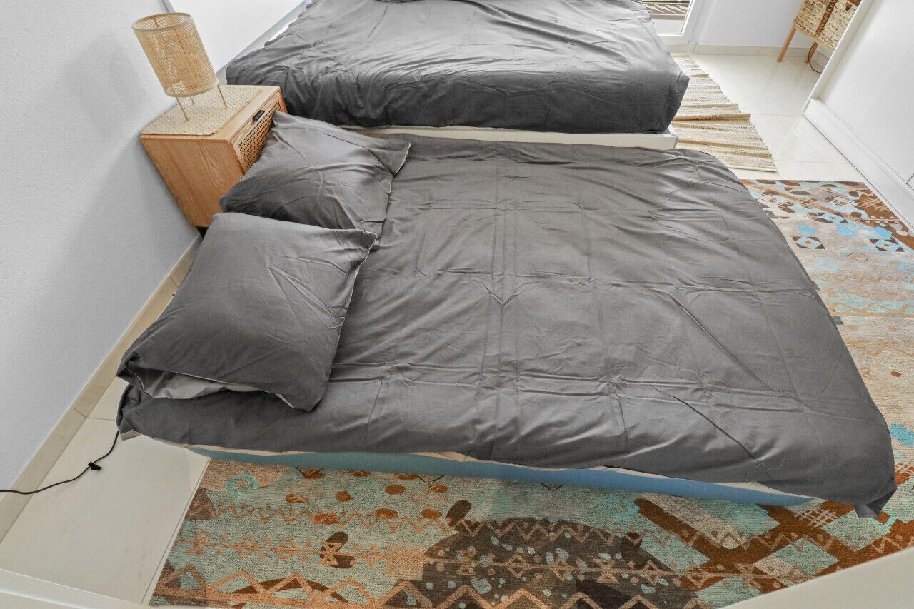 A bed with grey bedding and a wooden nightstand with a lamp.