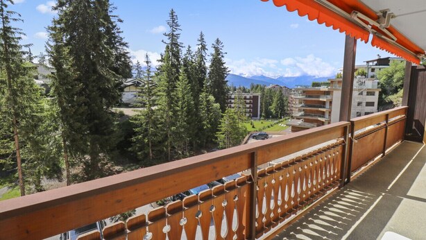 Een balkon met houten hekwerk en uitzicht op een bergachtig landschap met bomen en gebouwen.