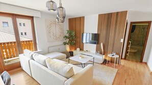 A modern living room with a sofa, coffee table, and TV, wooden flooring, and a balcony with a view. - Logement chaleureux au coeur de la station (Crans-Montana)