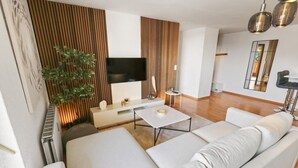 A modern living room with a large sofa, a flat-screen TV, a small side table, and a potted plant. - Logement chaleureux au coeur de la station (Crans-Montana)