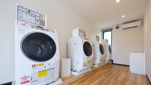A row of white front-loading washing machines in a clean, well-lit room with wooden flooring and an air conditioning unit on the wall. - HOTEL R9 The Yard Tomioka (Tomioka)