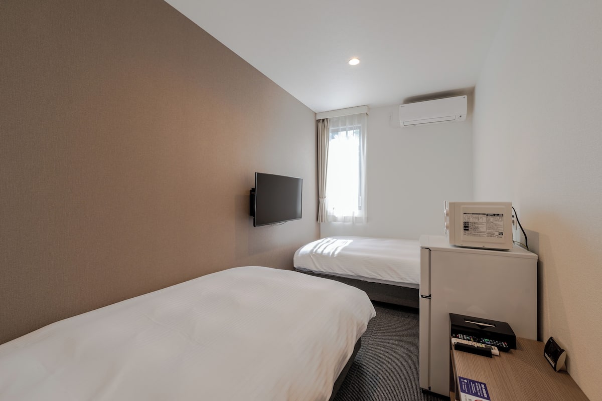 A hotel room with two single beds, a flat-screen TV mounted on the wall, a microwave on a small table, and a window with curtains.