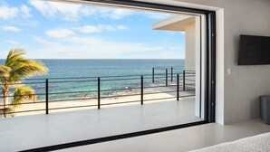 A modern room with a large sliding glass door leading to a balcony with a view of the ocean and palm trees. - Beachfront Estate with Full-Time Staff, Airport Transfer & Beach/Fitness Club Ac (San Jose Del Cabo)