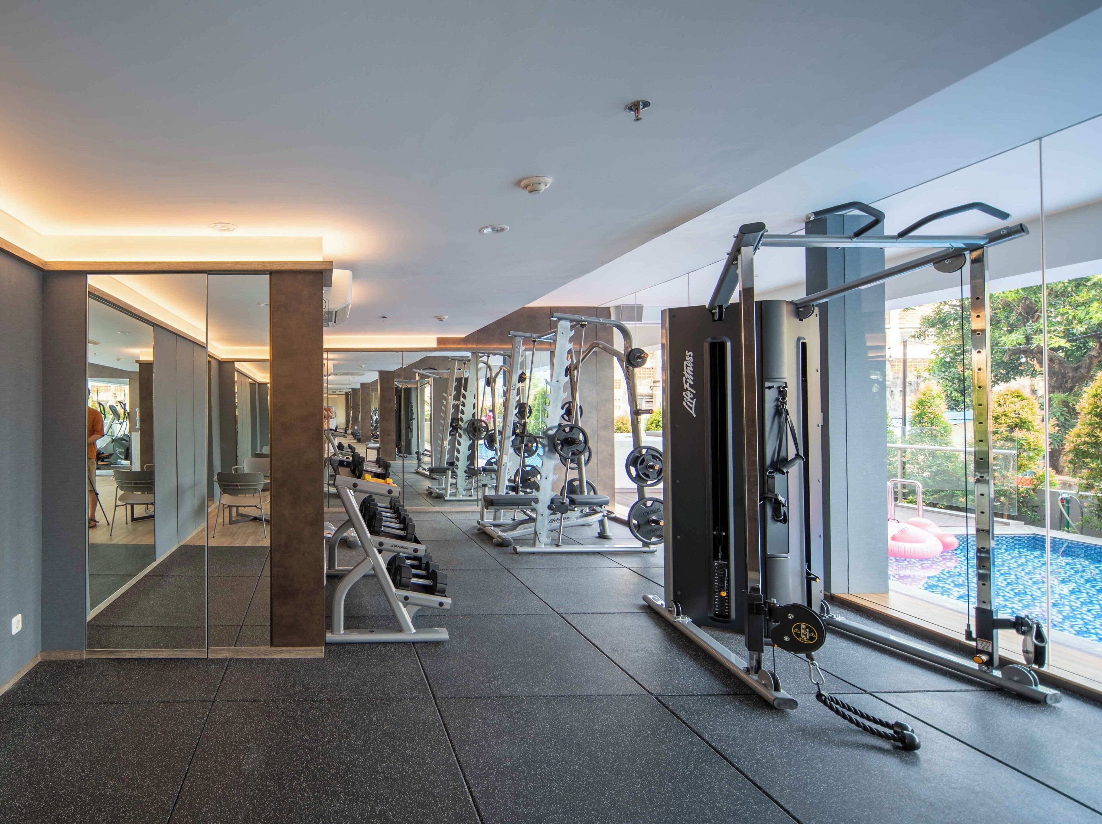Un gimnasio moderno con equipo de ejercicio variado, un espejo grande y vistas a una piscina.