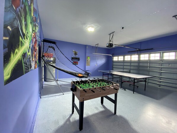 A room with a foosball table, a ping pong table, and a large window with a view of a garage.