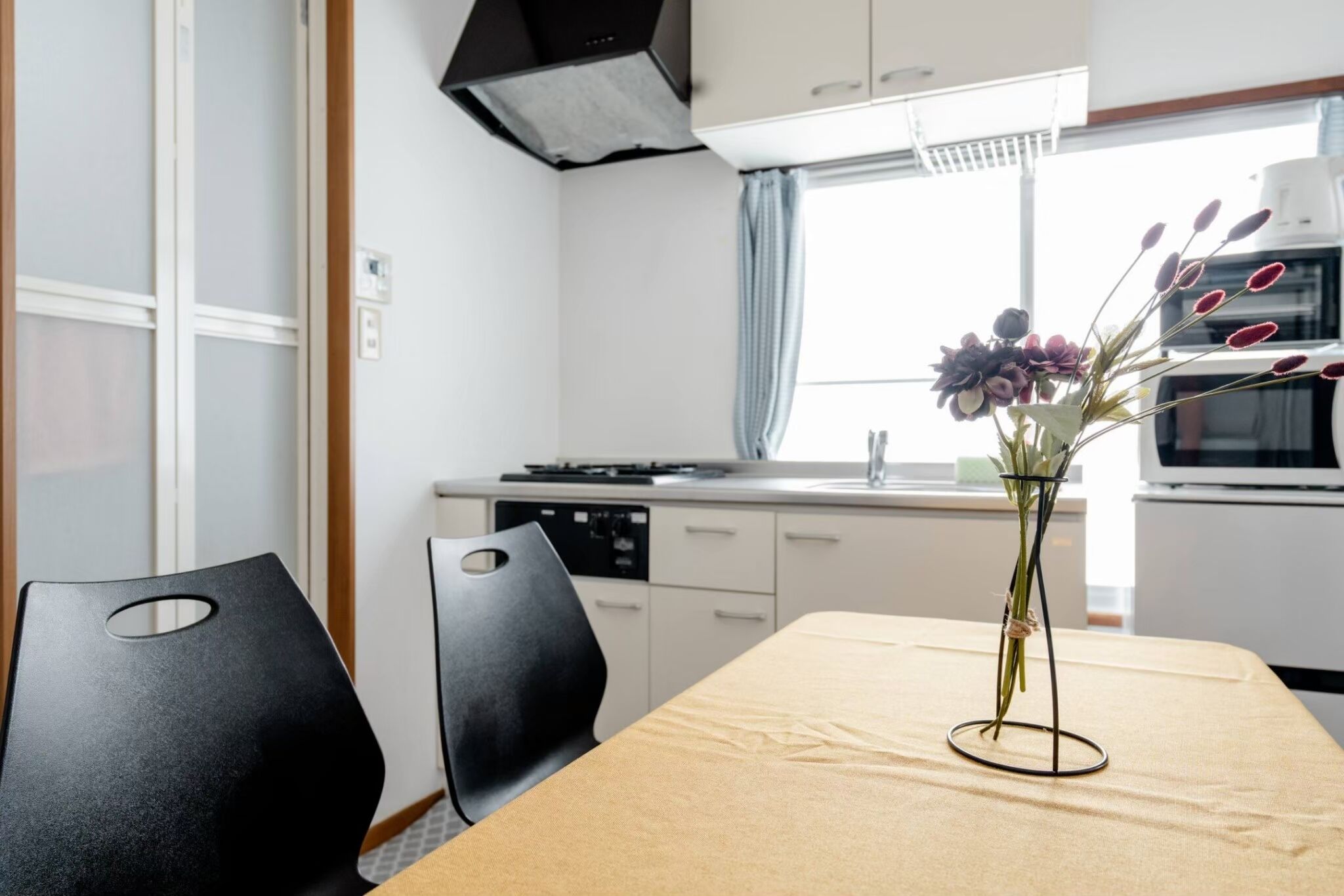 A modern kitchen with white cabinets, a stove, and a microwave. There is a dining table with a vase of flowers.