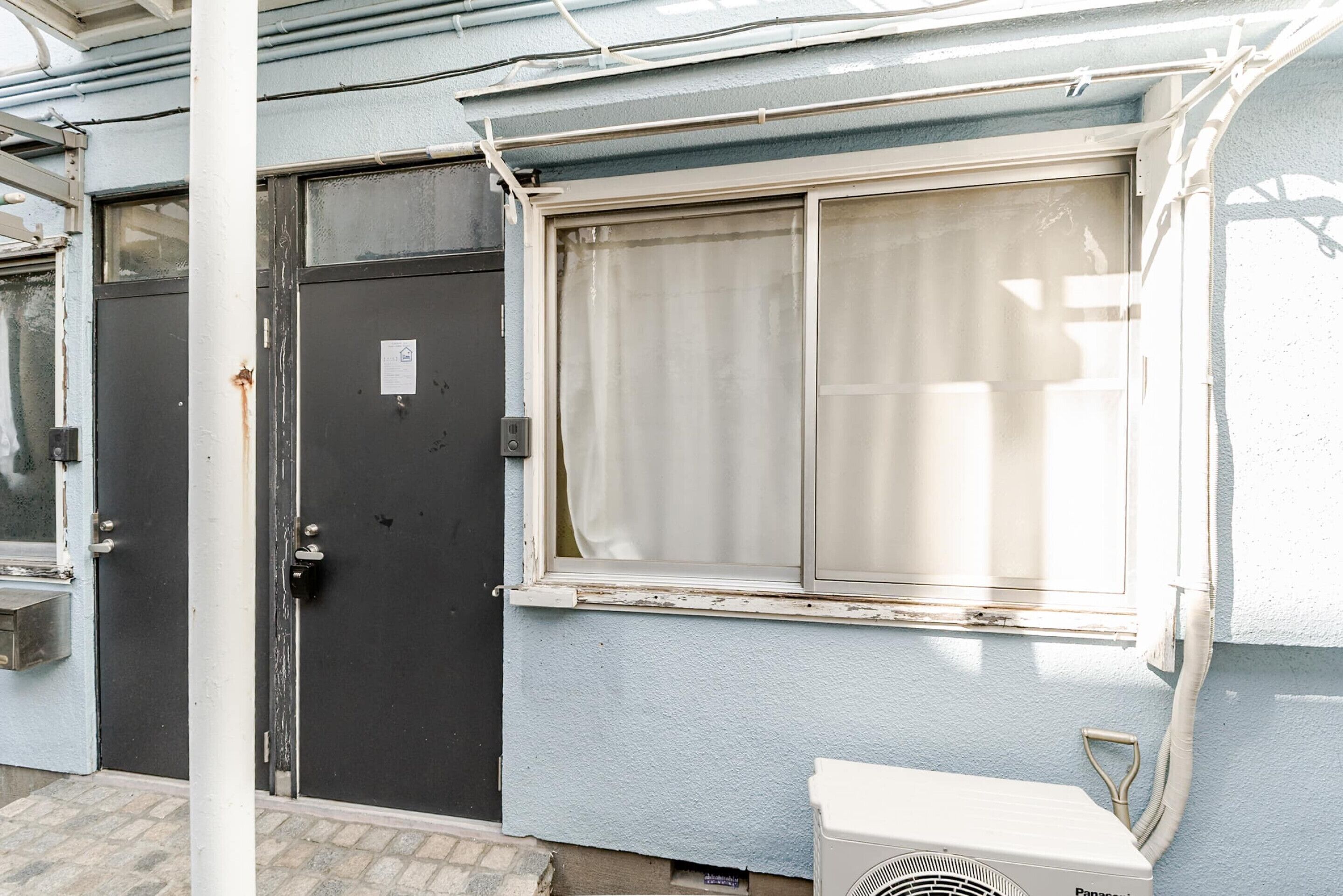 L’extérieur d’un bâtiment avec une porte noire fermée, une fenêtre avec un rideau et un appareil de climatisation blanc.