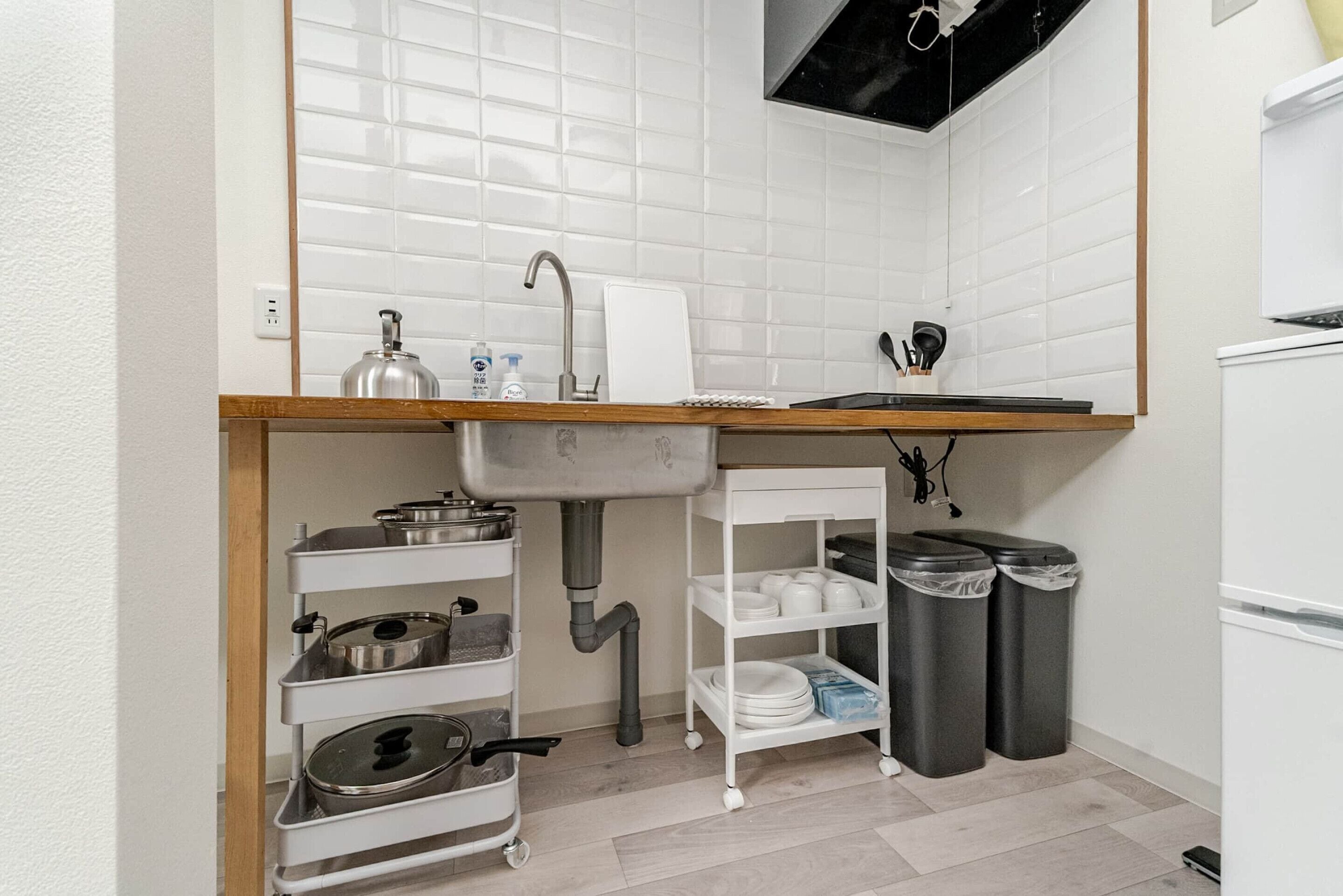 A compact kitchen with a sink, a white tiled backsplash, a wooden countertop, a white shelf unit, and two trash bins.