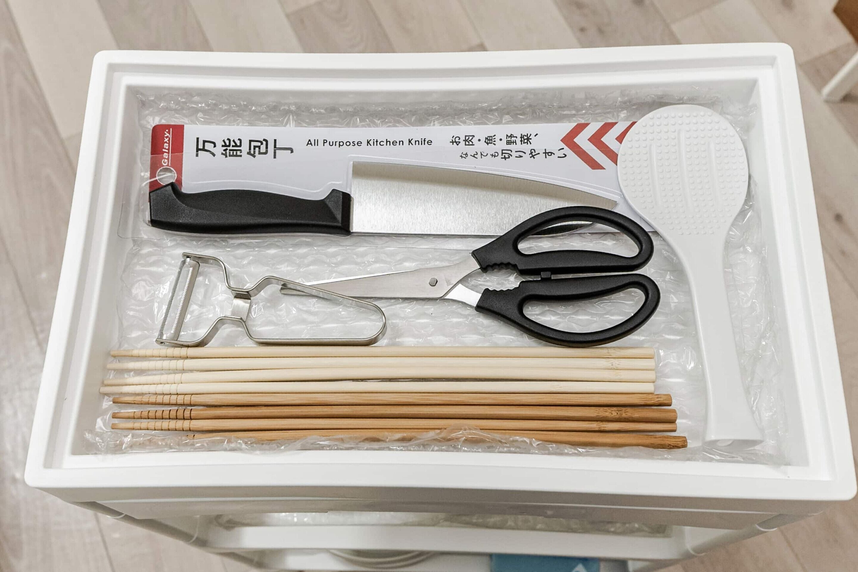 A white box containing a kitchen knife, scissors, a grater, and chopsticks.